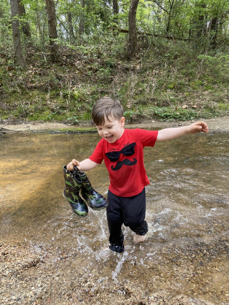 toddler playing in the water holding his rain boots in the forest